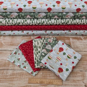 Peut inclure: Une pile de cinq pièces de tissu pliées présentant des motifs de montgolfières, de chalets d'hiver, de sapins et de fleurs rouges. Une règle en bois est placée sous le tissu, affichant des mesures en centimètres jusqu'à 38 cm.