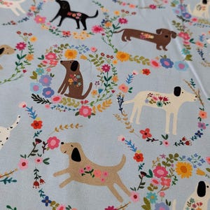 May include: Pale blue fabric with a repeating pattern of dogs, each decorated with floral motifs. The dogs are encircled by floral wreaths in pink, yellow, and blue. A delightful and whimsical design.
