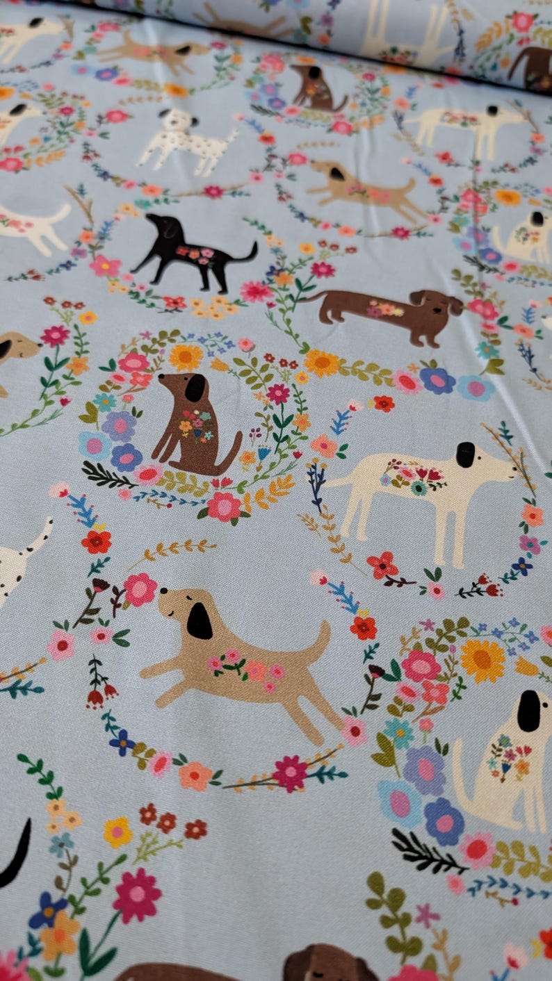 May include: Pale blue fabric with a repeating pattern of dogs, each decorated with floral motifs. The dogs are encircled by floral wreaths in pink, yellow, and blue. A delightful and whimsical design.