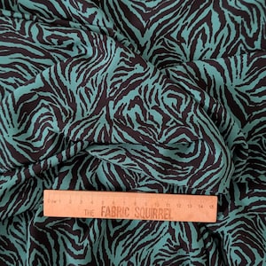 May include: Teal fabric featuring a black zebra stripe design. The fabric is folded, displaying the repeating pattern. A wooden ruler with centimetre markings and the text "THE FABRIC SQUIRREL" is placed on the fabric.