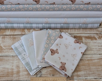 May include: A collection of five folded fabric swatches featuring patterns of brown teddy bears, stars, moons, buttons, and stripes in shades of grey, beige, and white. A wooden ruler is placed at the bottom showing 15 centimetres in length.