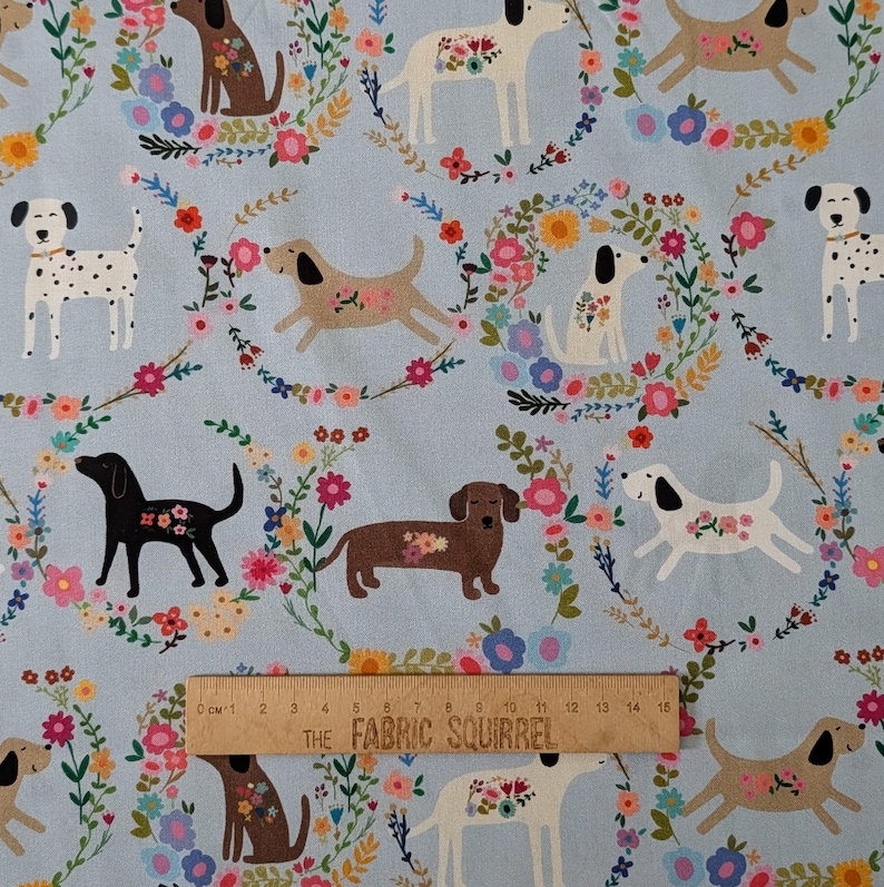 May include: Pale blue fabric with a repeating pattern of dogs and floral wreaths. The dogs are depicted in various breeds and colors, each with floral accents. A wooden ruler with metric measurements is visible at the bottom, with the text "THE FABRIC SQUIRREL".