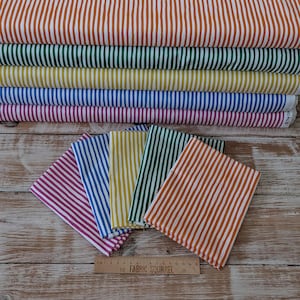 Könnte beinhalten: Eine Sammlung farbenfroher Stoffrollen und gefalteter Stoffquadrate mit Streifen. Der Stoff weist vertikale Streifen in verschiedenen Farben auf, darunter Orange, Grün, Gelb, Blau und Lila. Die gefalteten Quadrate sind auf einer Holzoberfläche mit einem Lineal angeordnet.