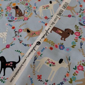 May include: Pale blue fabric featuring a repeating pattern of dogs and floral wreaths. The dogs are in various colours, including black, brown, and white. A white ribbon with text is across the fabric.