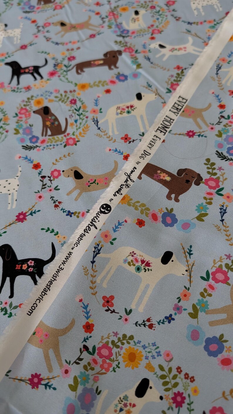 May include: Pale blue fabric featuring a repeating pattern of dogs and floral wreaths. The dogs are in various colours, including black, brown, and white. A white ribbon with text is across the fabric.