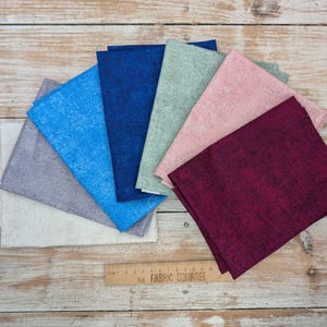 May include: A selection of fabric squares in shades of blue, grey, pink, and burgundy, displayed on a wooden surface. The fabrics have a textured finish. A ruler with the text "THE FABRIC SQUIRREL" is present.