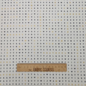 May include: Cream-coloured fabric featuring a repeating pattern of black and gold letters. The word "KINDNESS" is visible. A wooden ruler with centimetre markings and the text "THE FABRIC SQUIRREL" is placed on the fabric.
