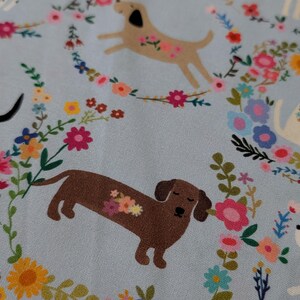 May include: Pale blue fabric featuring a repeating pattern of dogs, each decorated with flowers. The dogs are encircled by colourful floral wreaths, creating a cheerful and whimsical design.