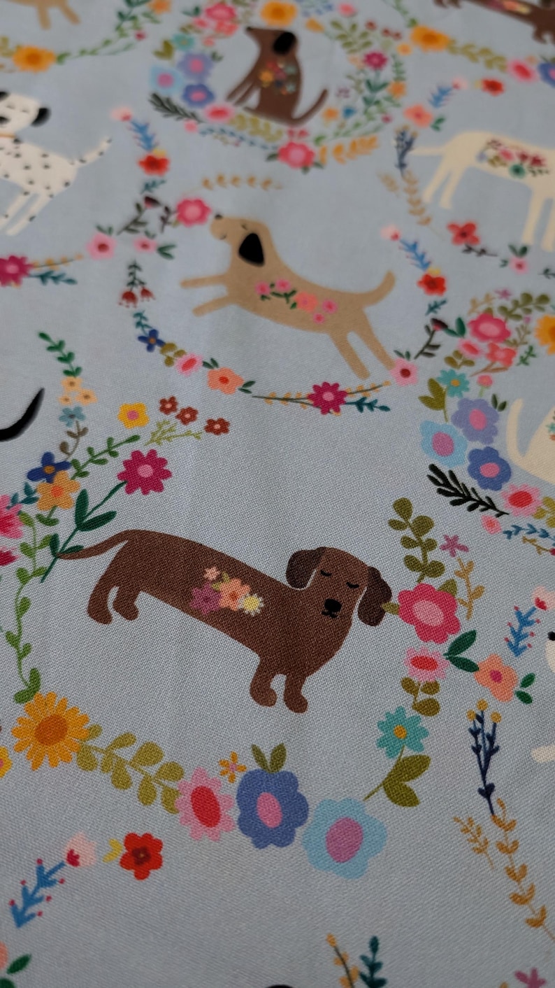 May include: Pale blue fabric featuring a repeating pattern of dogs, each decorated with flowers. The dogs are encircled by colourful floral wreaths, creating a cheerful and whimsical design.