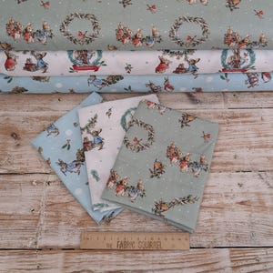 May include: Several rolls of patterned fabric with rabbit and wreath designs in blue, white, and green. The fabric is stacked on a wooden surface. A ruler with 'The Fabric Squirrel' printed on it is visible.