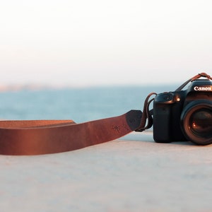 May include: A black Canon EOS camera with a brown leather camera strap. The camera is sitting on a concrete surface with a body of water in the background.