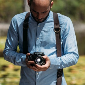 May include: A man wearing a light blue button-down shirt and brown leather camera straps is holding a black camera with a lens. The camera has the text "Mamiya 645" printed on it.