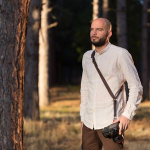 May include: A man with a beard wearing a white shirt and brown trousers stands in a forest. He is holding a camera with a brown leather strap.