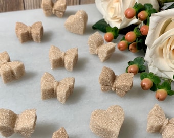 May include: A collection of glitter-covered, light brown sugar bow and heart-shaped decorations. The decorations are scattered on a white marble surface, with white roses and berries in the background.