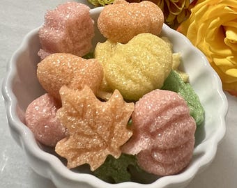 May include: A white scalloped bowl filled with assorted sugar-coated candies shaped like pumpkins and maple leaves in shades of pink, orange, yellow, and green. The textured, crystalline surface of each candy reflects light, creating a sparkling appearance.