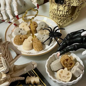 Może przedstawiać: Dwupoziomowa taca do serwowania z białej porcelany ze złotym wykończeniem zawiera kostki cukru w kształcie czaszek. Mała biała miseczka z falistym brzegiem zawiera kostki cukru w kształcie dyń. Czarny brokatowy pająk siedzi na tacy, otoczony figurkami szkieletów.