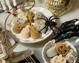 May include: A two-tiered white porcelain serving tray with gold trim holds sugar cubes shaped like skulls. A small white scalloped bowl contains sugar cubes shaped like pumpkins. A black glittery spider sits on the tray, surrounded by skeleton figures.