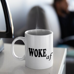 以下が含まれることがあります： 黒い文字で「WOKE af」と書かれた白いセラミック製のコーヒーカップ。カップから蒸気が上がっている。