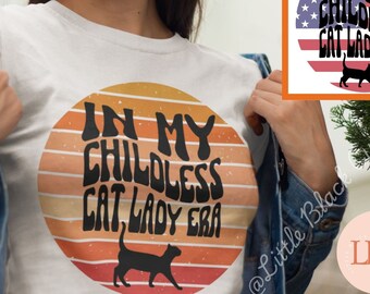 Op de afbeelding: Een wit T-shirt met een zwart en oranje grafisch ontwerp met de tekst "In My Childless Cat Lady Era" met een zwart katten silhouet. Het grafische ontwerp is een zonsondergang met oranje, gele en rode strepen.