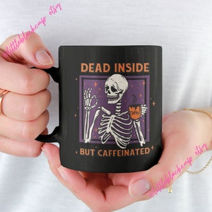 Pode incluir: Caneca de café preta com um gráfico branco de um esqueleto segurando uma xícara de café. O texto na caneca diz "Dead Inside + But Caffeinated +"