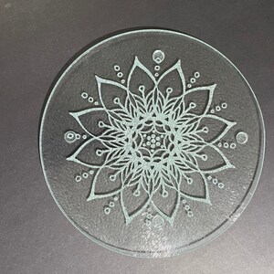 May include: A clear glass coaster with an etched mandala design. The mandala is white and intricate, with a circular pattern of flowers and leaves.