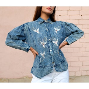 May include: A light blue denim shirt with embroidered floral designs. The shirt features a collared neckline, button-up front, and long sleeves with puffed shoulders. The floral embroidery includes white and red accents.