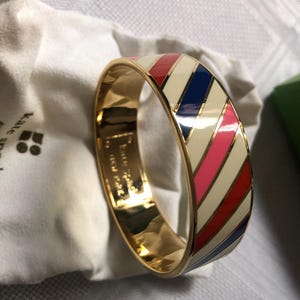 Könnte beinhalten: Ein goldenes Armreif-Armband mit einem weißen, roten, rosa und blauen Streifenmuster. Das Armband ist aus Metall und hat eine glänzende Oberfläche.