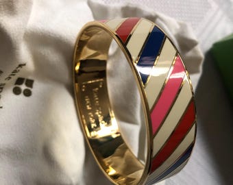 May include: A gold bangle bracelet with a white, red, pink and blue striped design. The bracelet is made of metal and has a glossy finish.