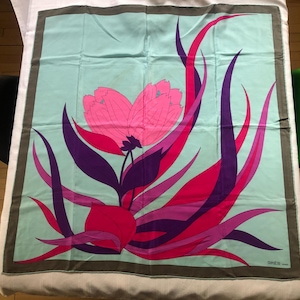 May include: Square silk scarf with a floral design. The scarf features a large pink flower with purple and red leaves against a light blue background. The border is a dark gray-brown color. The brand name "GRES" is in the bottom right corner.
