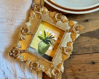 Peut inclure: Un petit cadre doré orné d'une peinture de fleurs blanches dans un vase vert. La peinture a un fond jaune. Le cadre est posé sur une surface en bois à côté d'un tissu crème à franges.