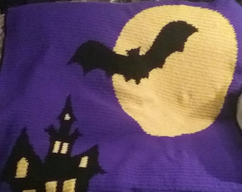 May include: A purple Halloween-themed blanket with a black silhouette of a bat flying in front of a yellow moon. A black haunted house and graveyard are also featured in the design.