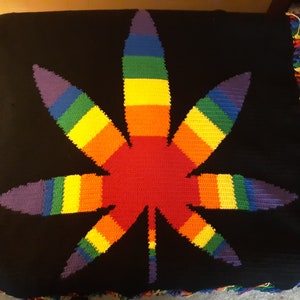 May include: A rainbow-colored crocheted blanket featuring a large marijuana leaf design. The blanket has a black background and a fringe border.