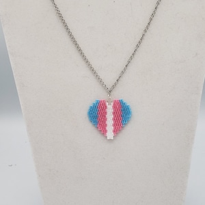 May include: A silver chain necklace with a heart-shaped pendant. The pendant is made of beads in the colors of the transgender pride flag: light blue, pink, and white.