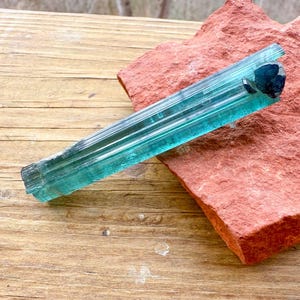 May include: A vibrant, blue-green tourmaline crystal rests on a rough, reddish-brown stone. The crystal is elongated with visible striations, and the stone has a natural, textured appearance. The background is a light brown wooden surface.