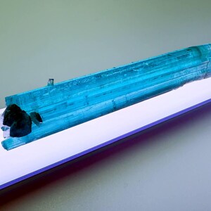 May include: A vibrant, elongated blue tourmaline crystal with a dark, irregular inclusion. The crystal is a striking shade of turquoise, displaying natural striations. The mineral specimen is presented on a white surface, creating a contrast that highlights its color and texture.