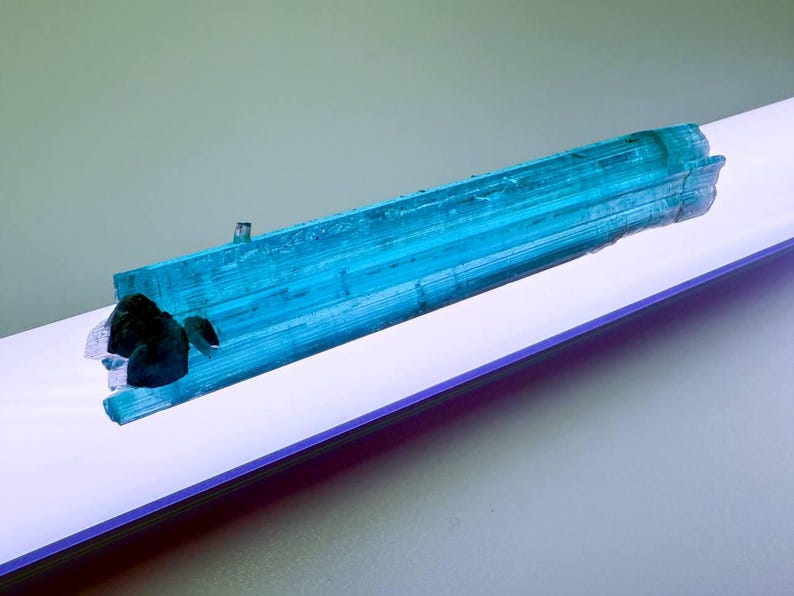 May include: A vibrant, elongated blue tourmaline crystal with a dark, irregular inclusion. The crystal is a striking shade of turquoise, displaying natural striations. The mineral specimen is presented on a white surface, creating a contrast that highlights its color and texture.