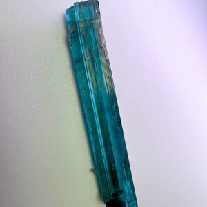May include: A vibrant, elongated teal crystal with vertical striations. The crystal has a translucent quality, allowing light to pass through, and features a dark, fractured base. The overall shape is a long, slender prism, showcasing natural geological formations.