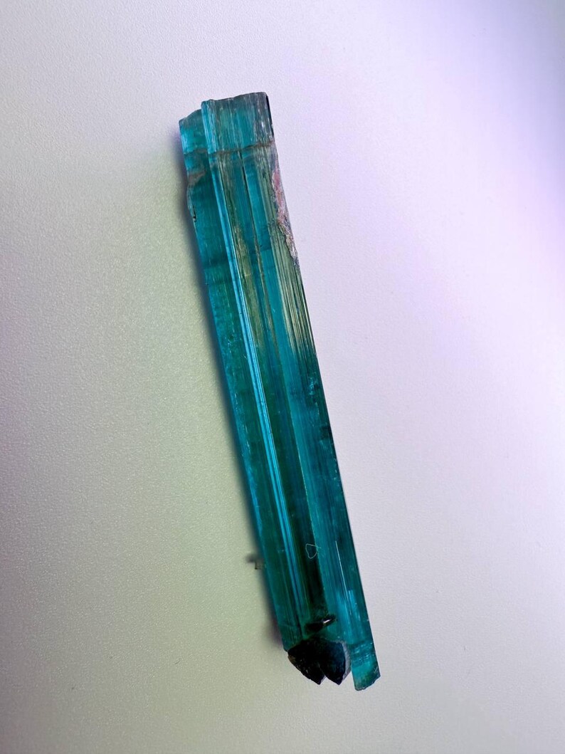 May include: A vibrant, elongated teal crystal with vertical striations. The crystal has a translucent quality, allowing light to pass through, and features a dark, fractured base. The overall shape is a long, slender prism, showcasing natural geological formations.