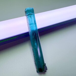 May include: A raw, elongated blue-green tourmaline crystal. The gemstone has a translucent quality with visible internal structures. The crystal is set against a white and blue background.
