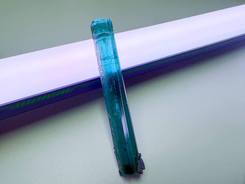 May include: A raw, elongated blue-green tourmaline crystal. The gemstone has a translucent quality with visible internal structures. The crystal is set against a white and blue background.