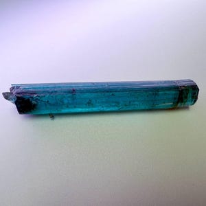 May include: A vibrant, elongated teal tourmaline crystal with a faceted, geometric shape. The gemstone displays a rich, translucent color gradient, transitioning from deep teal to a darker, almost black hue at the tip. The crystal is approximately 3 inches long.