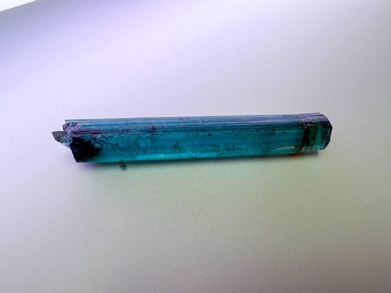 May include: A vibrant, elongated teal tourmaline crystal with a faceted, geometric shape. The gemstone displays a rich, translucent color gradient, transitioning from deep teal to a darker, almost black hue at the tip. The crystal is approximately 3 inches long.