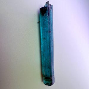 May include: A raw, elongated teal tourmaline crystal with a dark, almost black, base and top. The crystal is vertically oriented, displaying a translucent quality with visible striations. The overall shape is a long, slender prism.