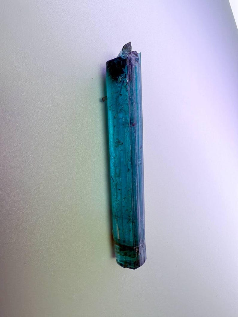 May include: A raw, elongated teal tourmaline crystal with a dark, almost black, base and top. The crystal is vertically oriented, displaying a translucent quality with visible striations. The overall shape is a long, slender prism.
