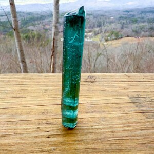 May include: A tall, translucent, teal-colored crystal stands upright on a wooden surface. The crystal has a layered appearance with bands of lighter and darker teal. The background features a blurred view of trees and a distant landscape.