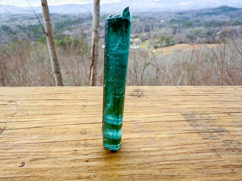 May include: A tall, translucent, teal-colored crystal stands upright on a wooden surface. The crystal has a layered appearance with bands of lighter and darker teal. The background features a blurred view of trees and a distant landscape.