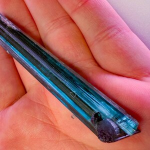May include: A blue and green tourmaline crystal is held in a hand. The crystal has a layered appearance with bands of dark green, teal, and blue. The crystal's ends are dark and rough. The background is a pinkish hue.