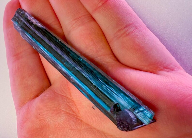May include: A blue and green tourmaline crystal is held in a hand. The crystal has a layered appearance with bands of dark green, teal, and blue. The crystal's ends are dark and rough. The background is a pinkish hue.