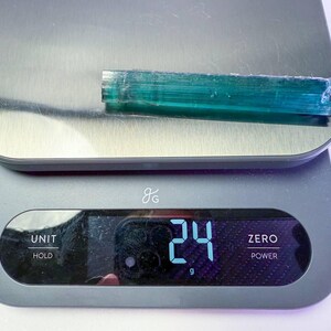 May include: A teal-colored, elongated crystal is placed on a digital scale. The scale displays a weight of 24 grams. The crystal is wrapped in clear plastic. The scale is gray with a silver top.