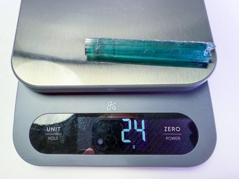 May include: A teal-colored, elongated crystal is placed on a digital scale. The scale displays a weight of 24 grams. The crystal is wrapped in clear plastic. The scale is gray with a silver top.